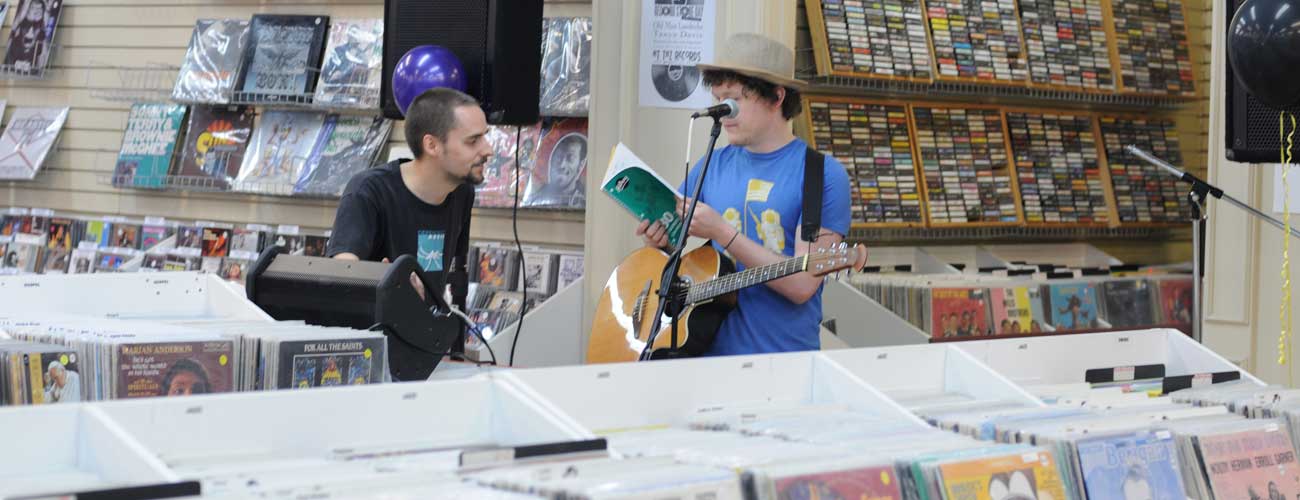 Image contains a photo of two people talking with each other in the background of a record store. One guy has an acoustic guitar and is standing behind a microphone while opening a notebook.